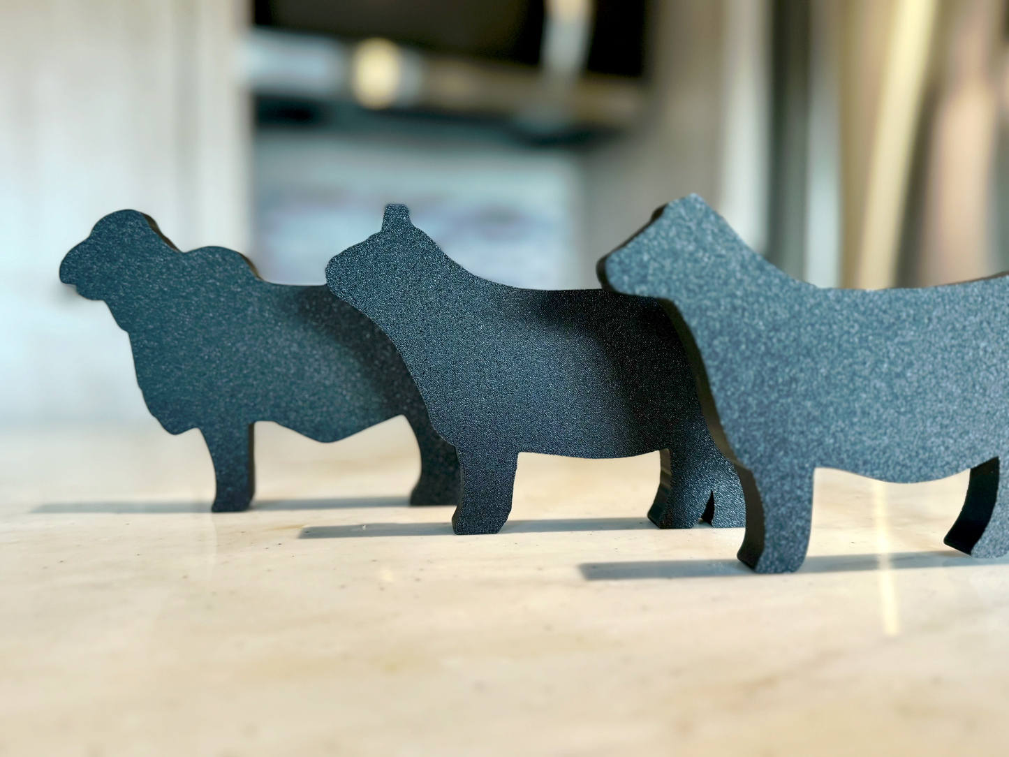 Three farm animal-shaped tabletop decorations made of PLA plastic, featuring a Charolais cow, a Western Highland cow, and a gray Brahman cow. They are arranged in a row on a light-colored countertop, with a modern kitchen in the background, including a stove and a tiled backsplash.