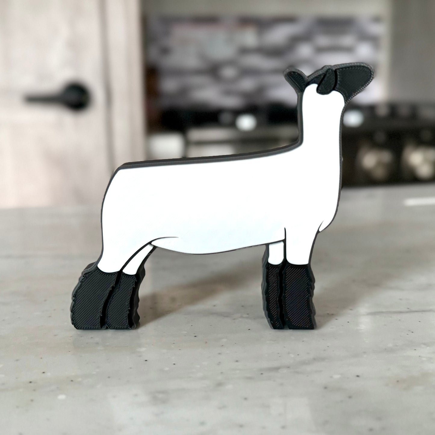 A 3D-printed market lamb figurine featuring a sleek white body, black head, and fluffy black legs, standing on a smooth countertop. The modern, minimalist design showcases clean lines and a show-ready stance, making it a perfect decorative piece for livestock enthusiasts.