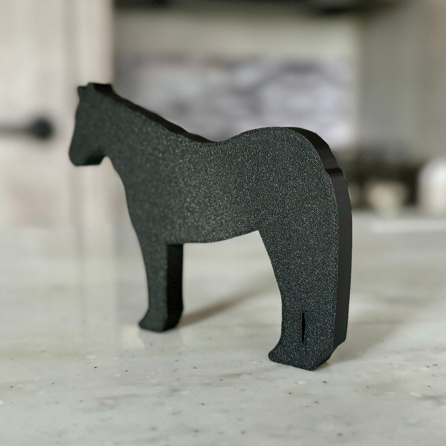 A horse-shaped tabletop decoration made of PLA plastic, featuring a brown body with black outlines and white accents on the hooves and face. The horse stands on a light-colored countertop, with a modern kitchen in the background, including a stove and tiled backsplash.