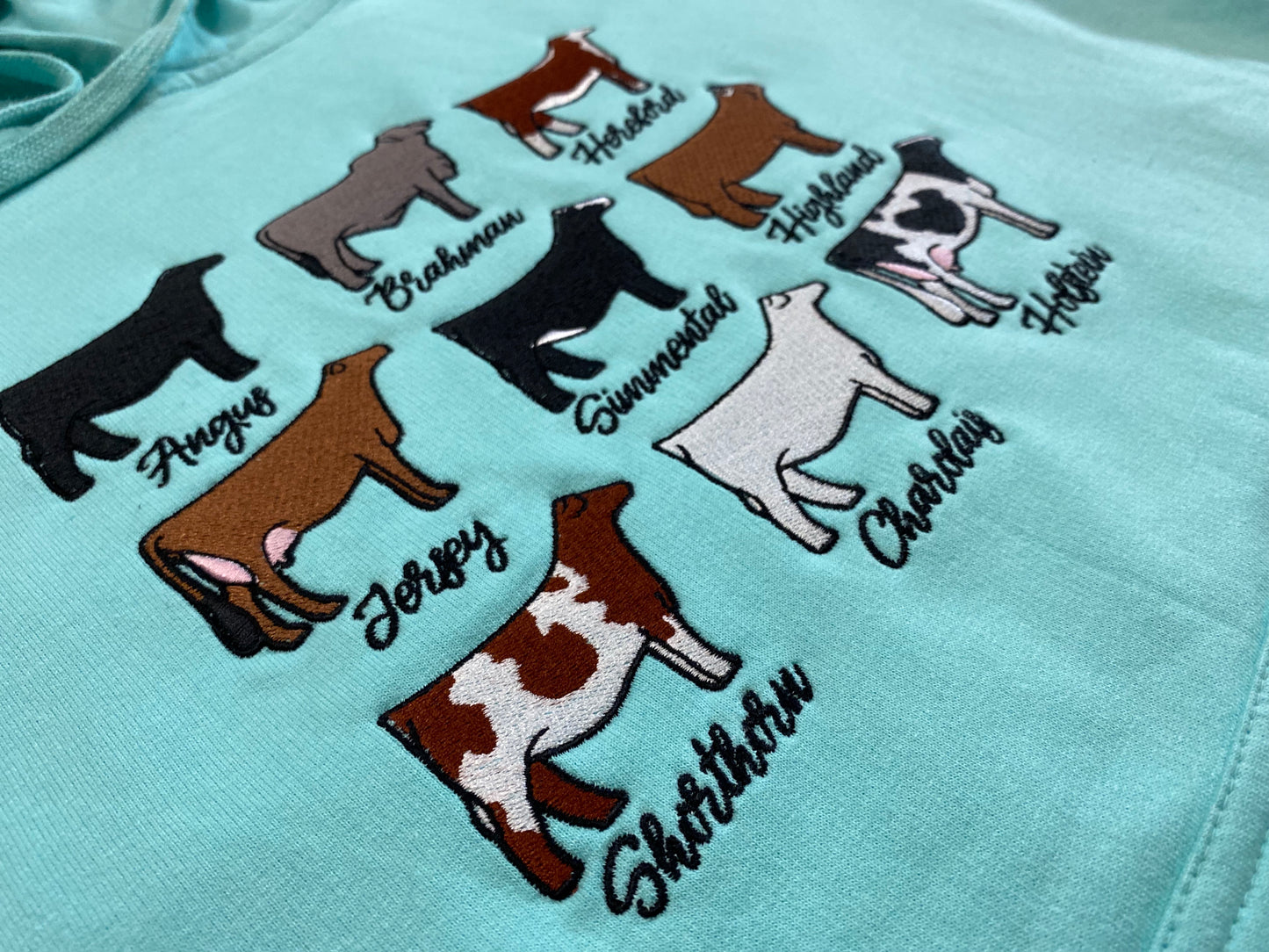 Turquoise hoodie featuring a graphic design of nine different cattle breeds, including Angus, Brahman, Hereford, Jersey, Simmental, Highland, Shorthorn, Charolais, and Holstein, each labeled with their breed name. The hoodie has a front pocket and a drawstring hood, laid flat on a brown and white cowhide background.