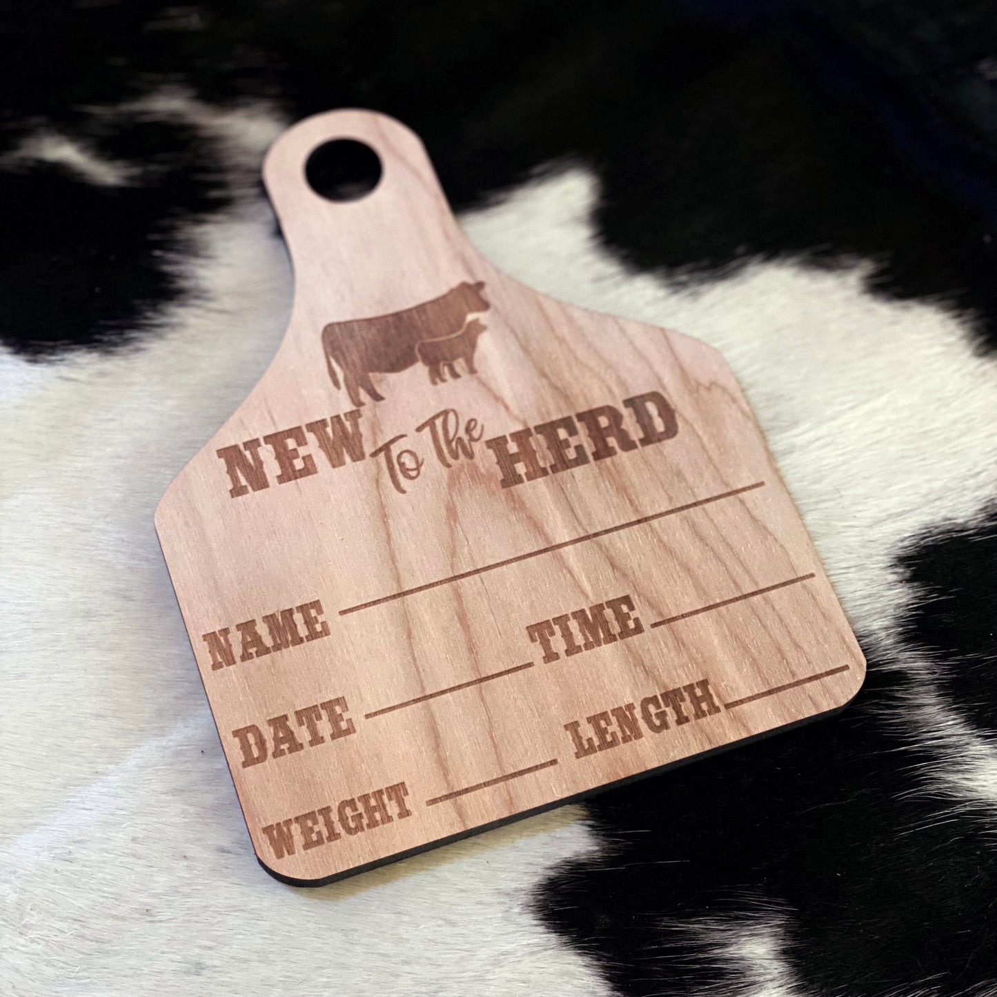 A wooden birth announcement sign shaped like an ear tag with "New to the Herd" engraved at the top, along with a cow and calf silhouette. The sign features engraved sections for filling in a baby's name, date, time of birth, weight, and length. It is placed on a black and white cowhide background, giving it a rustic, farm-themed aesthetic.