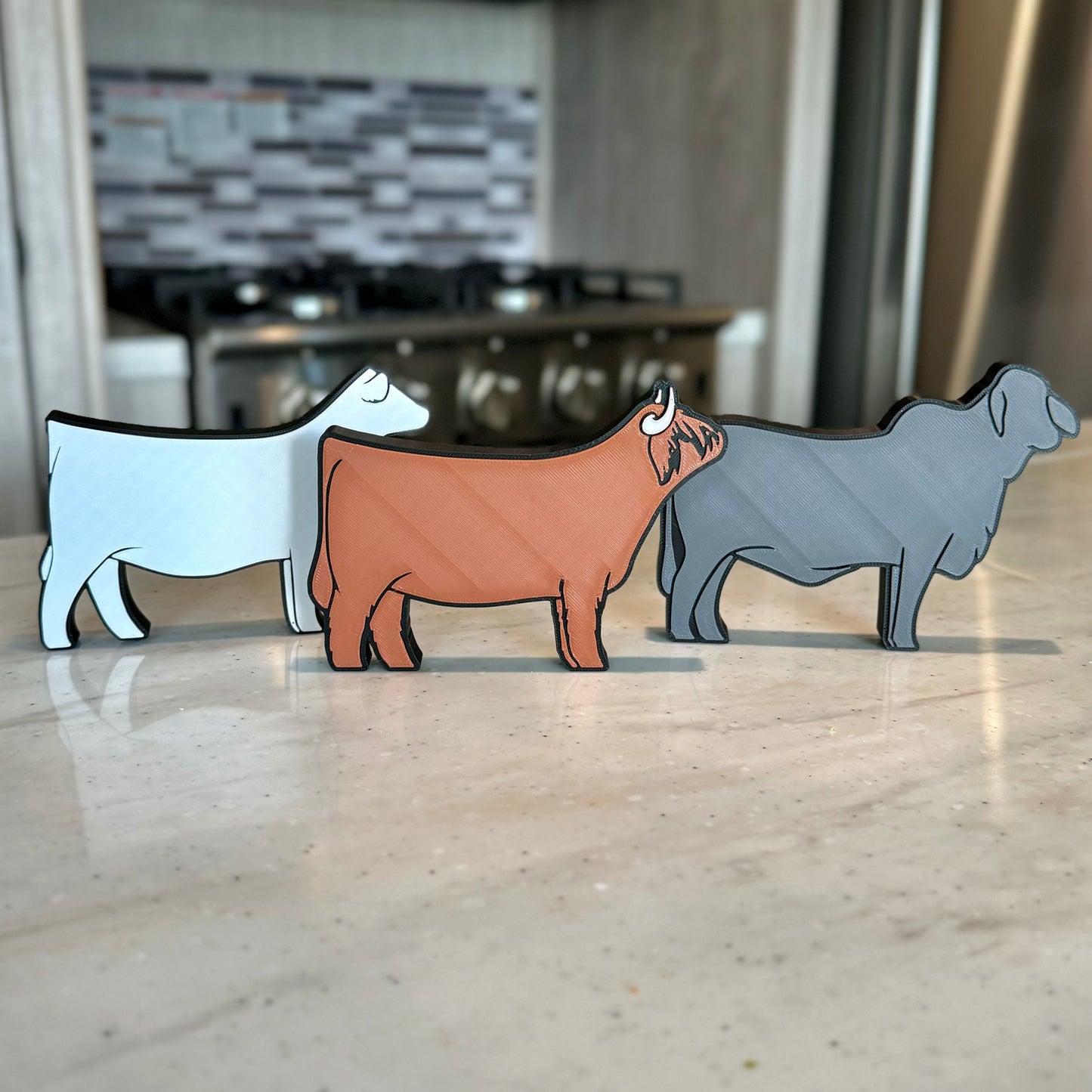 Three farm animal-shaped tabletop decorations made of PLA plastic, featuring a Charolais cow, a Western Highland cow, and a gray Brahman cow. They are arranged in a row on a light-colored countertop, with a modern kitchen in the background, including a stove and a tiled backsplash.