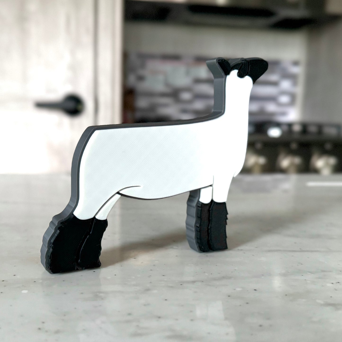 A 3D-printed market lamb figurine featuring a sleek white body, black head, and fluffy black legs, standing on a smooth countertop. The modern, minimalist design showcases clean lines and a show-ready stance, making it a perfect decorative piece for livestock enthusiasts.