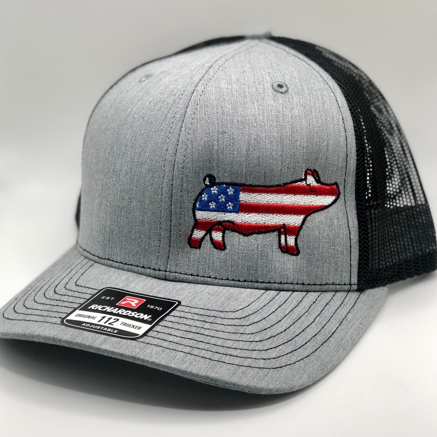 Gray Richardson 112 trucker hat with a black mesh back, featuring an embroidered pig silhouette filled with an American flag design. The pig embroidery has red and white stripes with a blue star section on its body. The hat has a structured front panel, a curved brim, and an adjustable snapback closure. A Richardson brand sticker is visible on the brim. The hat is displayed against a neutral background.