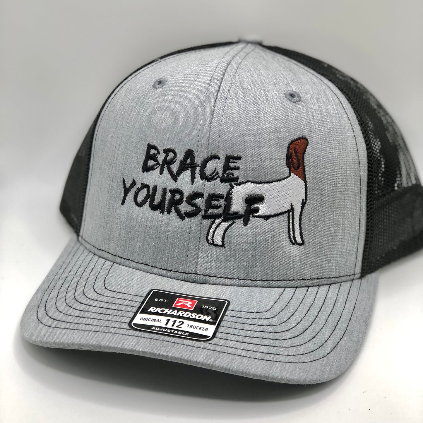 A Richardson 112 trucker hat with a grey front panel and black mesh back. The front features an embroidered design with the phrase "Brace Yourself" in bold black lettering, along with an image of a Boer show goat in a bracing stance. The hat has an adjustable snapback closure and a structured fit, perfect for livestock enthusiasts, show goat owners, and farm life lovers.