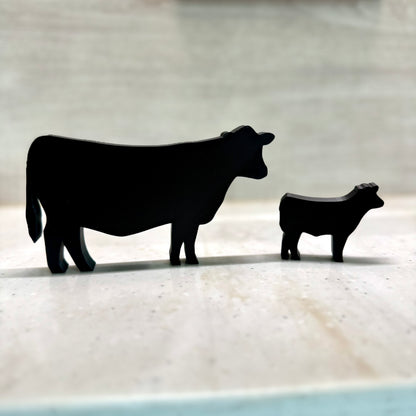 A 3D-printed black silhouette of a cow and calf standing on a light-colored surface. The minimalist design features smooth edges and a matte finish, creating a modern yet rustic farm-inspired decor piece. The background is softly blurred, emphasizing the sleek, simplistic shapes of the animals