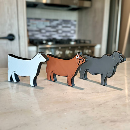 Three farm animal-shaped tabletop decorations made of PLA plastic, featuring a Charolais cow, a Western Highland cow, and a gray Brahman cow. They are arranged in a row on a light-colored countertop, with a modern kitchen in the background, including a stove and a tiled backsplash.