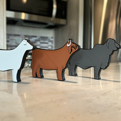 Three farm animal-shaped tabletop decorations made of PLA plastic, featuring a Charolais cow, a Western Highland cow, and a gray Brahman cow. They are arranged in a row on a light-colored countertop, with a modern kitchen in the background, including a stove and a tiled backsplash.