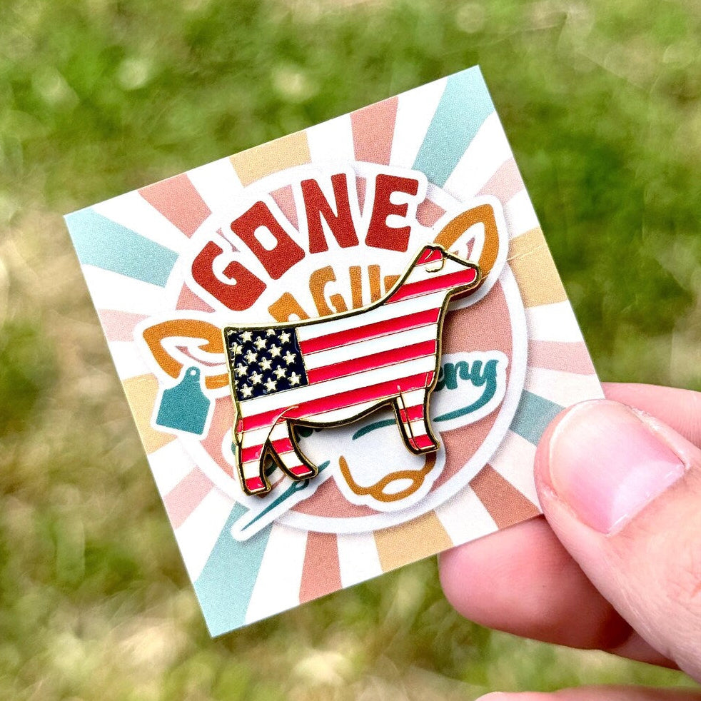 Cow shaped pin with American flag design on a colorful card held by a hand against a grassy background