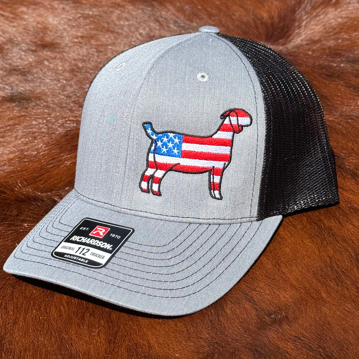 A Richardson 112 trucker hat in heather gray and black mesh, featuring an embroidered goat graphic with an American flag pattern on the front, resting on a brown fur surface.