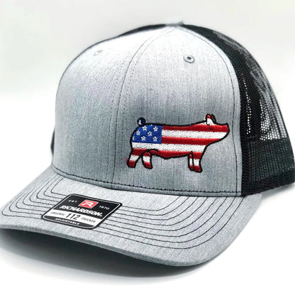 Gray Richardson 112 trucker hat with a black mesh back, featuring an embroidered pig silhouette filled with an American flag design. The pig embroidery has red and white stripes with a blue star section on its body. The hat has a structured front panel, a curved brim, and an adjustable snapback closure. A Richardson brand sticker is visible on the brim. The hat is displayed against a neutral background.
