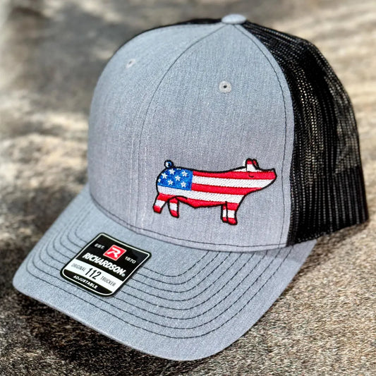 Gray Richardson 112 trucker hat with a black mesh back, featuring an embroidered pig silhouette filled with an American flag design. The pig embroidery has red and white stripes with a blue star section on its body. The hat has a structured front panel, a curved brim, and an adjustable snapback closure. A Richardson brand sticker is visible on the brim. The hat is displayed against a neutral background.