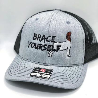 A Richardson 112 trucker hat with a grey front panel and black mesh back. The front features an embroidered design with the phrase "Brace Yourself" in bold black lettering, along with an image of a Boer show goat in a bracing stance. The hat has an adjustable snapback closure and a structured fit, perfect for livestock enthusiasts, show goat owners, and farm life lovers.