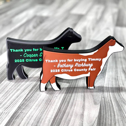 Two custom cow-shaped plaques are displayed on a wood-patterned surface. The front plaque is brown and white with the text, “Thank you for buying Timmy – Bethany Richburg, 2025 Citrus County Fair,” in white. The back plaque is black with green text reading, “Thank you for buying Mr. T – Cooper S…, 2025 Citrus County Fair.” These plaques are personalized thank-you gifts typically used in livestock shows or county fairs.