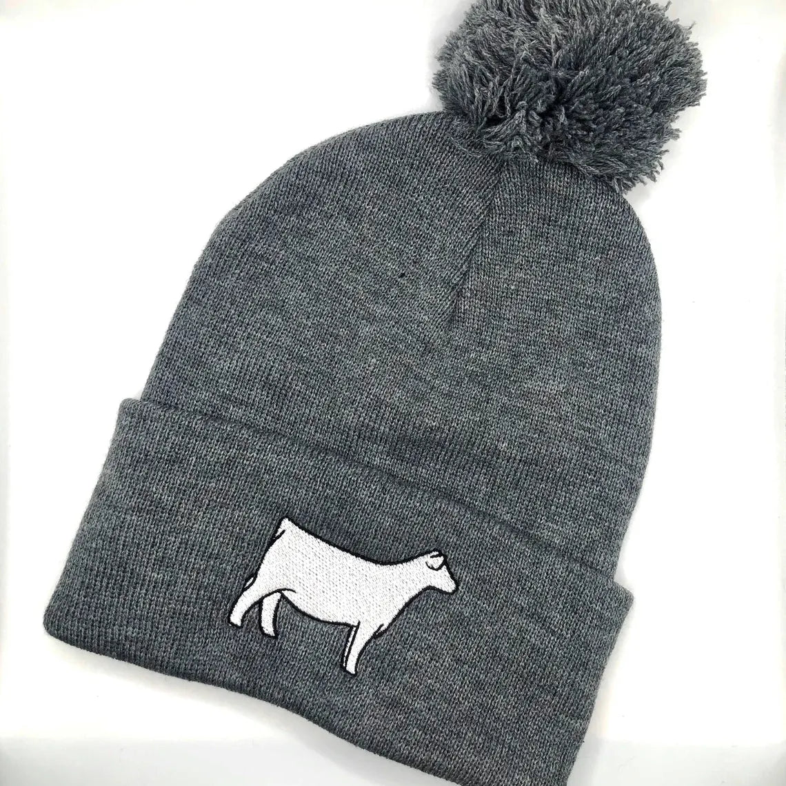 A gray knit beanie with a pom-pom on top, featuring an embroidered white Charolais cow design on the folded cuff. The beanie is laid flat against a white background.