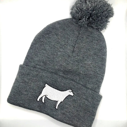 A gray knit beanie with a pom-pom on top, featuring an embroidered white Charolais cow design on the folded cuff. The beanie is laid flat against a white background.