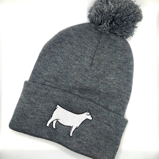 A gray knit beanie with a pom-pom on top, featuring an embroidered white Charolais cow design on the folded cuff. The beanie is laid flat against a white background.