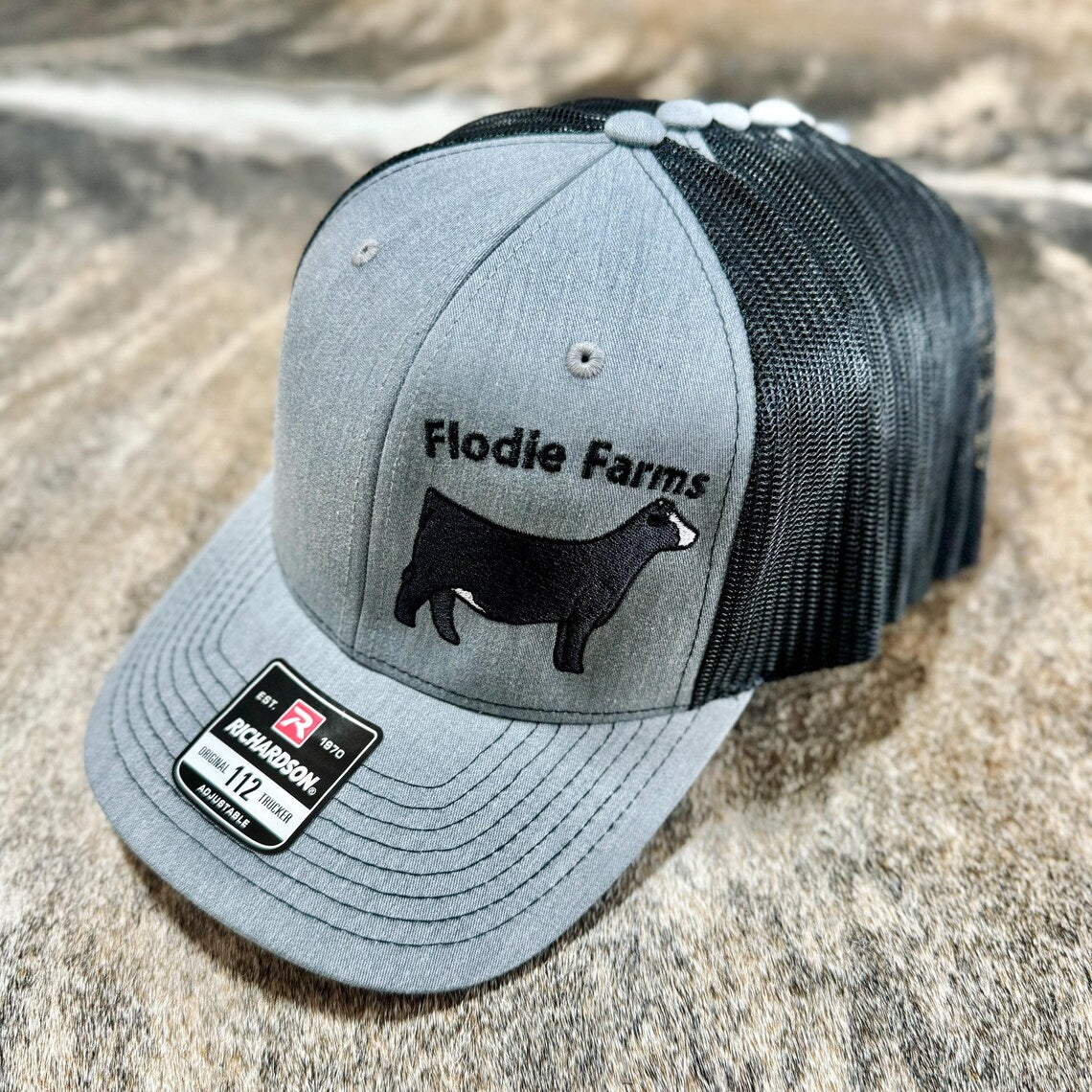 Gray and black cap customized with the farm name 'Flodie Farms' featuring a graphic of a Simmental heifer below.