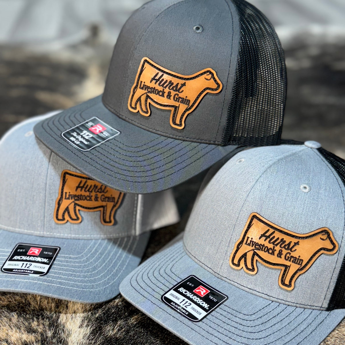 Two gray and black hats with 'Hurst Livestock & Grain' engraved on cow shaped leather patches on a blurred background.