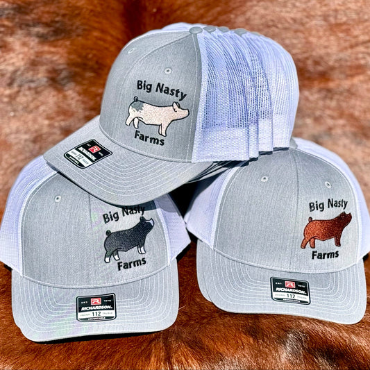Three gray and white baseball caps with 'Big Nasty Farms' branding and animal illustrations on a brown fur background.