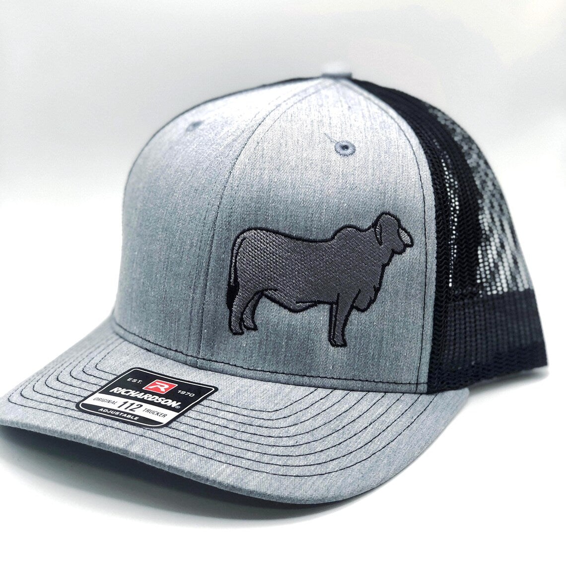 A custom embroidered trucker hat featuring a detailed silhouette of a Brahman cow on the front panel. Available in three color combinations: grey with black mesh, grey with white mesh, and charcoal with black mesh. The hat has a breathable mesh back and an adjustable snapback closure, offering a comfortable fit for both adult and youth sizes. Perfect for livestock enthusiasts, ranchers, and cattle show participants.