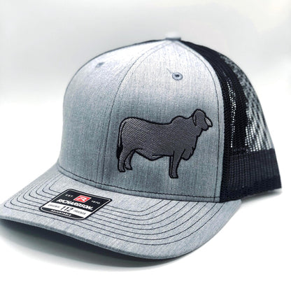 A custom embroidered trucker hat featuring a detailed silhouette of a Brahman cow on the front panel. Available in three color combinations: grey with black mesh, grey with white mesh, and charcoal with black mesh. The hat has a breathable mesh back and an adjustable snapback closure, offering a comfortable fit for both adult and youth sizes. Perfect for livestock enthusiasts, ranchers, and cattle show participants.