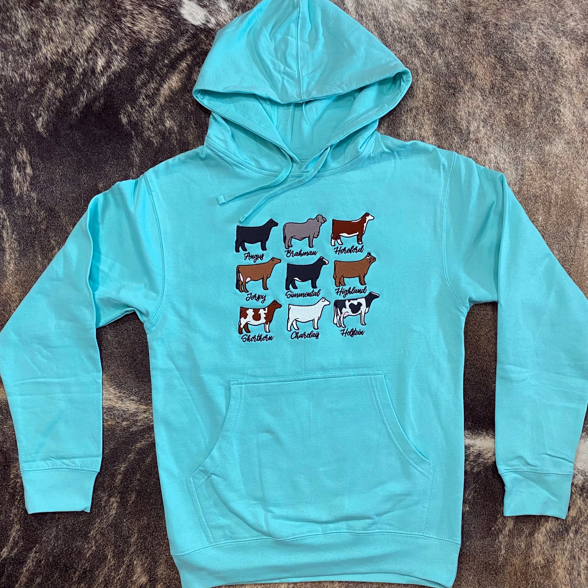 Turquoise hoodie featuring a graphic design of nine different cattle breeds, including Angus, Brahman, Hereford, Jersey, Simmental, Highland, Shorthorn, Charolais, and Holstein, each labeled with their breed name. The hoodie has a front pocket and a drawstring hood, laid flat on a brown and white cowhide background.