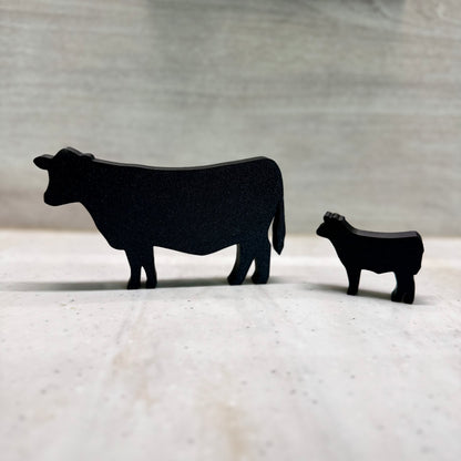 A 3D-printed black silhouette of a cow and calf standing on a light-colored surface. The minimalist design features smooth edges and a matte finish, creating a modern yet rustic farm-inspired decor piece. The background is softly blurred, emphasizing the sleek, simplistic shapes of the animals