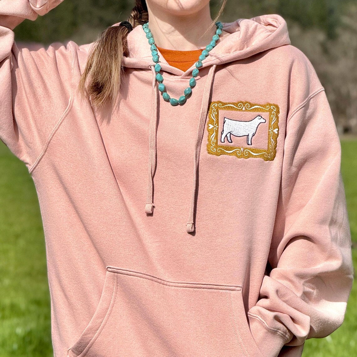 Person wearing a pink hoodie with a show heifer frame design outdoors.