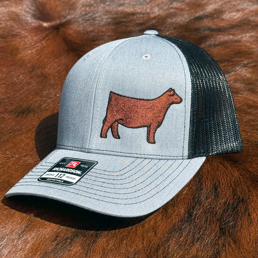 A light gray heather trucker cap with an embroidered red Angus cow on the front, displayed on a brown fur surface.