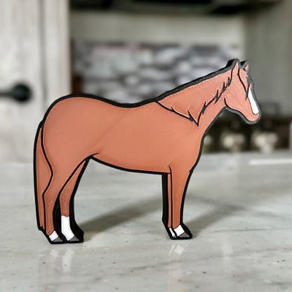 A horse-shaped tabletop decoration made of PLA plastic, featuring a brown body with black outlines and white accents on the hooves and face. The horse stands on a light-colored countertop, with a modern kitchen in the background, including a stove and tiled backsplash.