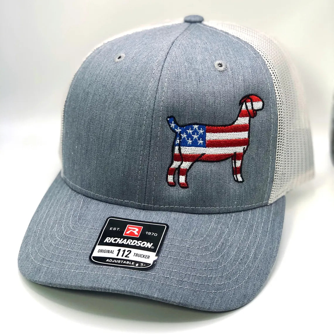 A Richardson 112 trucker hat featuring an embroidered silhouette of a goat filled with an American flag design, incorporating red, white, and blue stars and stripes. The hat is available in three color combinations: grey with black mesh, grey with white mesh, and charcoal with black mesh. It has a breathable mesh back and an adjustable snapback closure, making it a comfortable and stylish choice for farm enthusiasts and those looking to showcase their patriotic spirit.