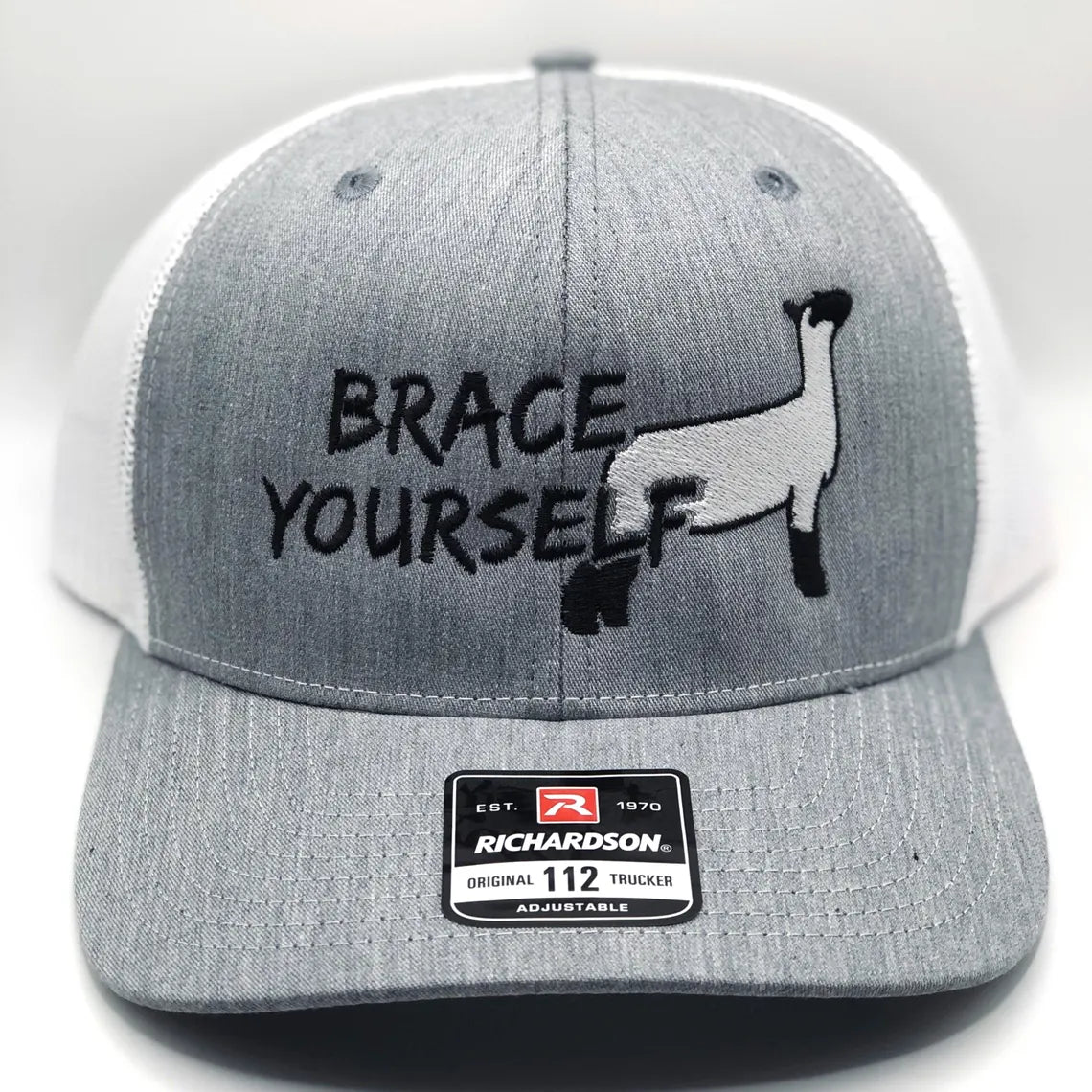 Gray and white Richardson 112 trucker hat featuring an embroidered design of a show sheep in black and white, along with the phrase "Brace Yourself" in bold black lettering. The hat has a structured front panel, a curved brim, and an adjustable mesh back for breathability and comfort.