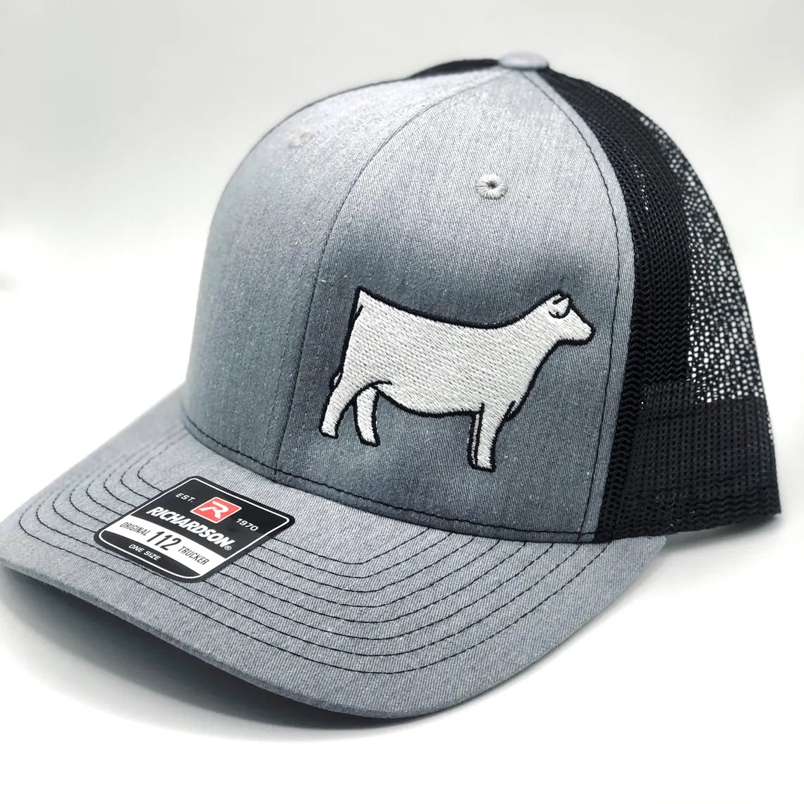 A gray and black Richardson 112 trucker hat with an embroidered white Charolais cow design on the front panel. The hat features a structured front, a curved brim, and a black mesh back for ventilation. A brand sticker is visible on the brim.