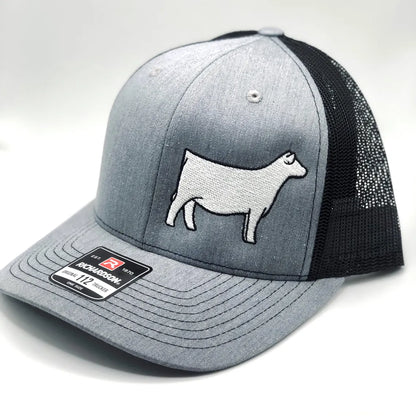 A gray and black Richardson 112 trucker hat with an embroidered white Charolais cow design on the front panel. The hat features a structured front, a curved brim, and a black mesh back for ventilation. A brand sticker is visible on the brim.