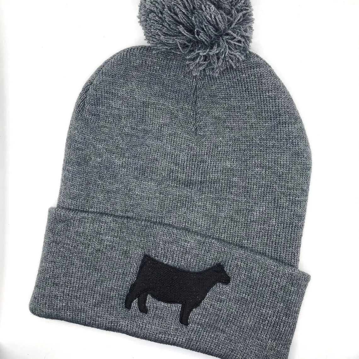 A dark grey knit beanie with a pom-pom on top, featuring a black embroidered show steer or Angus heifer silhouette on the folded cuff. The beanie is made of soft, ribbed knit fabric, providing a cozy and stylish look.