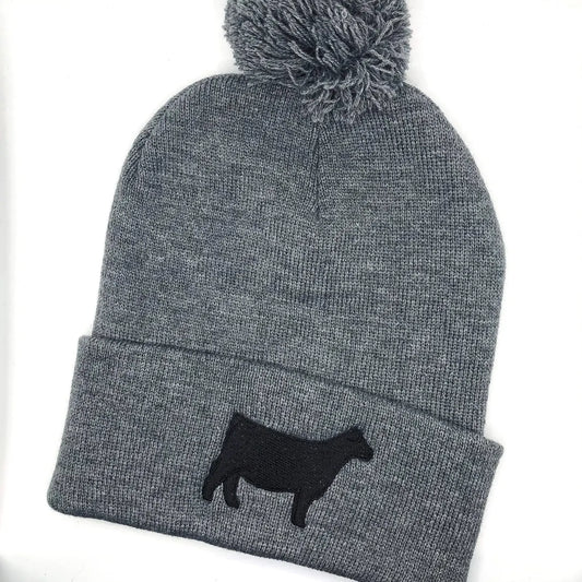 A dark grey knit beanie with a pom-pom on top, featuring a black embroidered show steer or Angus heifer silhouette on the folded cuff. The beanie is made of soft, ribbed knit fabric, providing a cozy and stylish look.