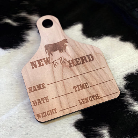 A wooden birth announcement sign shaped like an ear tag with "New to the Herd" engraved at the top, along with a cow and calf silhouette. The sign features engraved sections for filling in a baby&#39;s name, date, time of birth, weight, and length. It is placed on a black and white cowhide background, giving it a rustic, farm-themed aesthetic.