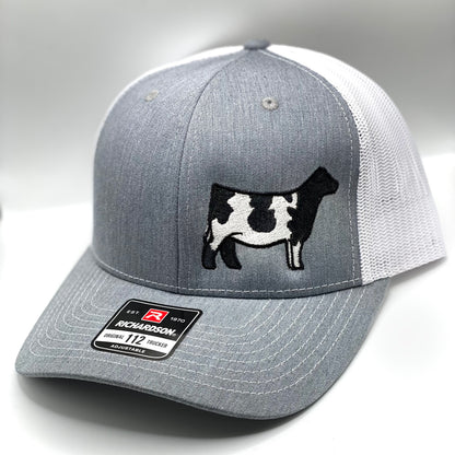 A custom embroidered trucker hat featuring a detailed Shorthorn cow silhouette on the front panel. Available in three color combinations: grey with black mesh, grey with white mesh, and charcoal with black mesh. Designed with a breathable mesh back and an adjustable snapback closure, this hat comes in both adult and youth sizes, making it a perfect fit for livestock enthusiasts of all ages.