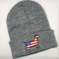 A grey knit beanie with an embroidered patch of a goat silhouette featuring an American flag design, with red and white stripes and a blue field with white stars. The beanie has a folded cuff and is laid flat on a white background.