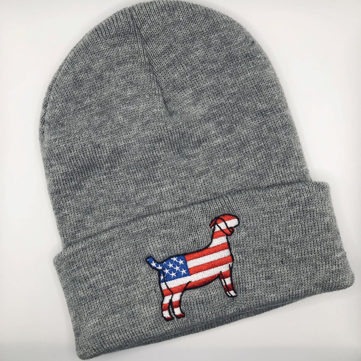 A grey knit beanie with an embroidered patch of a goat silhouette featuring an American flag design, with red and white stripes and a blue field with white stars. The beanie has a folded cuff and is laid flat on a white background.