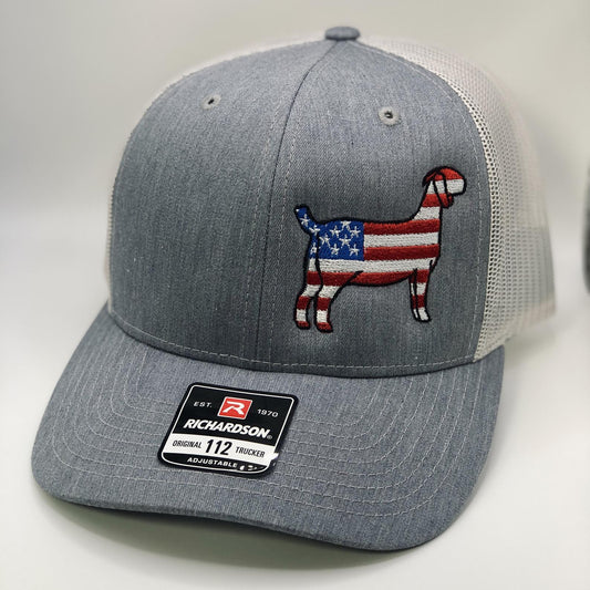 A Richardson 112 trucker hat featuring an embroidered silhouette of a goat filled with an American flag design, incorporating red, white, and blue stars and stripes. The hat is available in three color combinations: grey with black mesh, grey with white mesh, and charcoal with black mesh. It has a breathable mesh back and an adjustable snapback closure, making it a comfortable and stylish choice for farm enthusiasts and those looking to showcase their patriotic spirit.