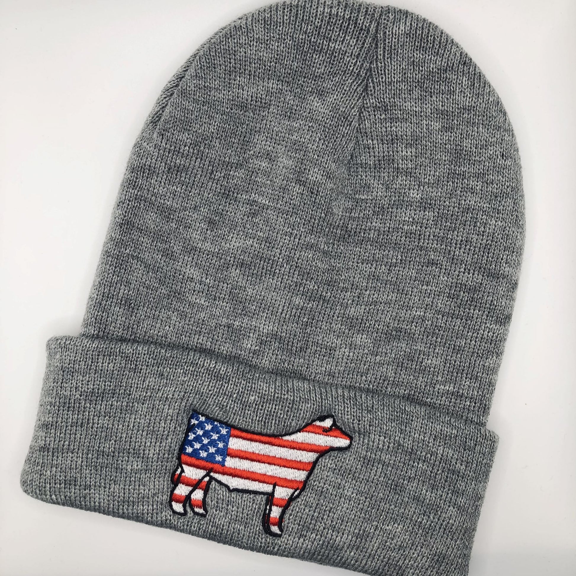 A gray knit beanie with an embroidered show steer patch featuring the American flag design. The steer silhouette is outlined in black, with red and white stripes on the body and a blue field with white stars on the front. The beanie has a folded cuff and is laid flat on a white background.