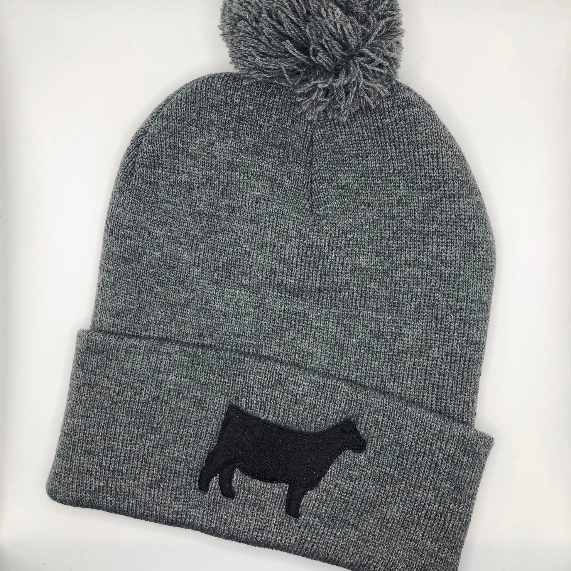 A dark grey knit beanie with a pom-pom on top, featuring a black embroidered show steer or Angus heifer silhouette on the folded cuff. The beanie is made of soft, ribbed knit fabric, providing a cozy and stylish look.