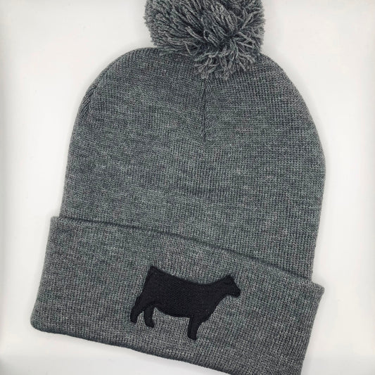A dark grey knit beanie with a pom-pom on top, featuring a black embroidered show steer or Angus heifer silhouette on the folded cuff. The beanie is made of soft, ribbed knit fabric, providing a cozy and stylish look.