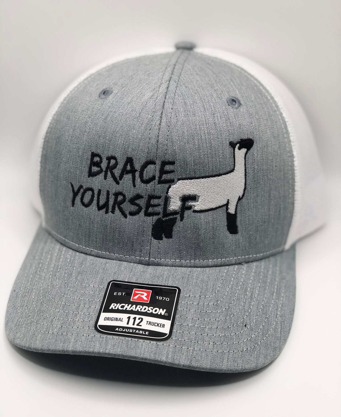 Gray and white Richardson 112 trucker hat featuring an embroidered design of a show sheep in black and white, along with the phrase "Brace Yourself" in bold black lettering. The hat has a structured front panel, a curved brim, and an adjustable mesh back for breathability and comfort.