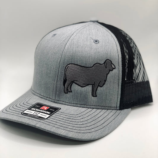 A custom embroidered trucker hat featuring a detailed silhouette of a Brahman cow on the front panel. Available in three color combinations: grey with black mesh, grey with white mesh, and charcoal with black mesh. The hat has a breathable mesh back and an adjustable snapback closure, offering a comfortable fit for both adult and youth sizes. Perfect for livestock enthusiasts, ranchers, and cattle show participants.