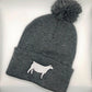 A gray knit beanie with a pom-pom on top, featuring an embroidered white Charolais cow design on the folded cuff. The beanie is laid flat against a white background.