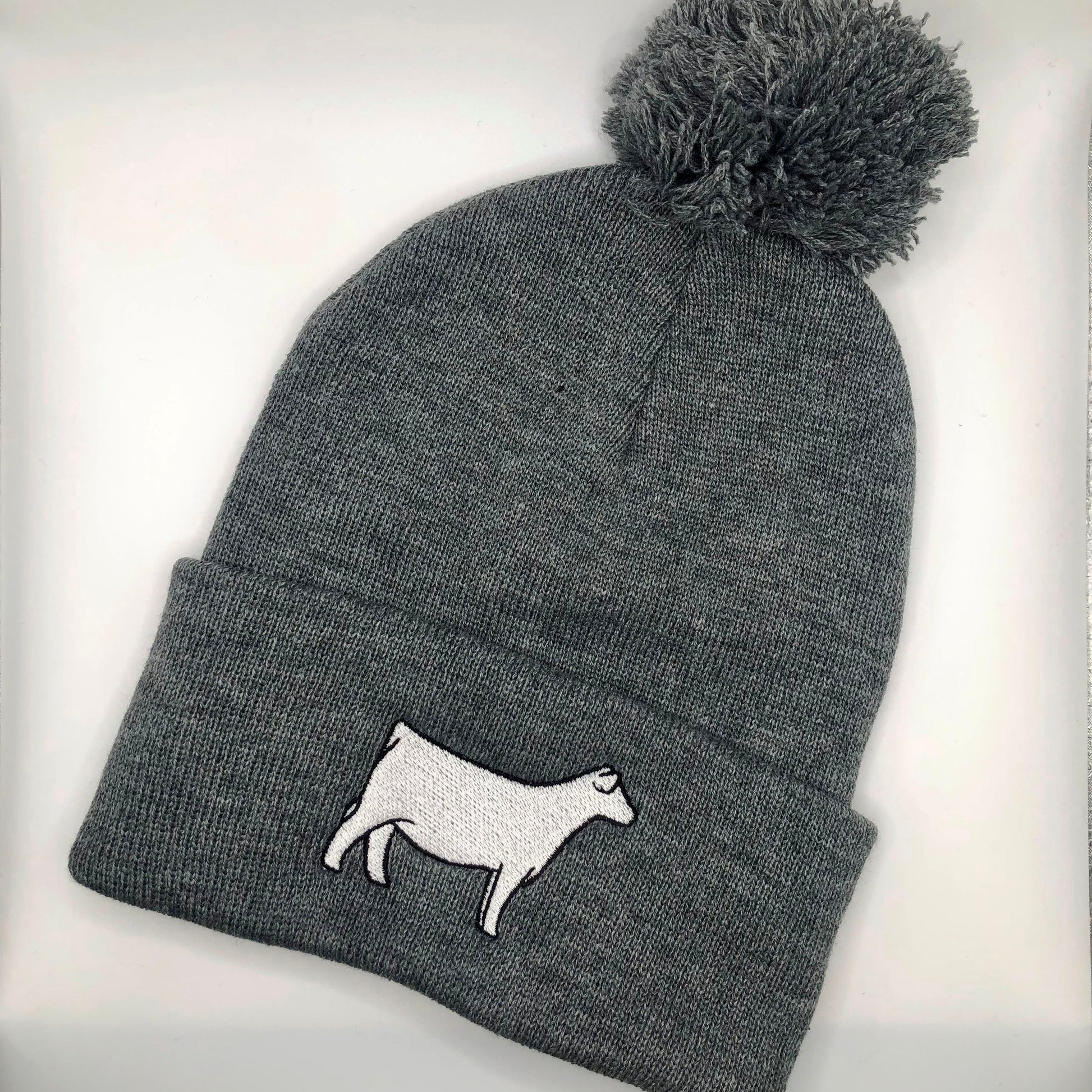 A gray knit beanie with a pom-pom on top, featuring an embroidered white Charolais cow design on the folded cuff. The beanie is laid flat against a white background.