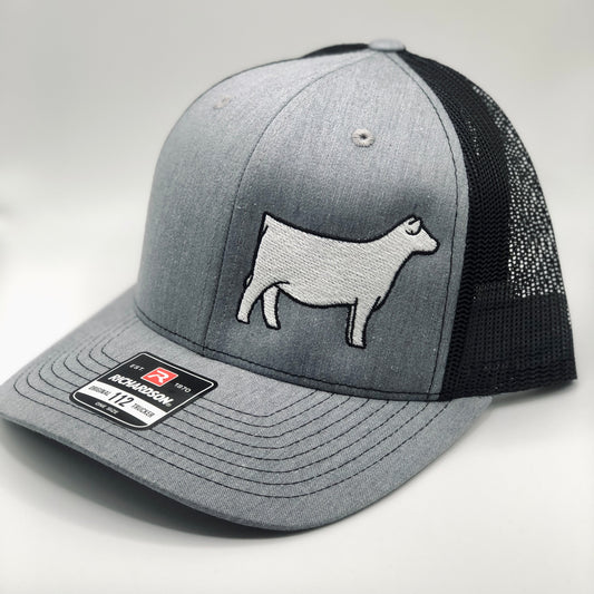 A gray and black Richardson 112 trucker hat with an embroidered white Charolais cow design on the front panel. The hat features a structured front, a curved brim, and a black mesh back for ventilation. A brand sticker is visible on the brim.